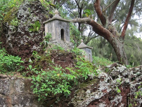 Two small stone structures nestled among lush greenery and rocky terrain, with trees in the background.