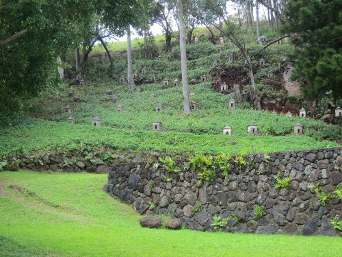 A hillside covered in greenery, dotted with small stone structures, surrounded by trees and a stone wall.
