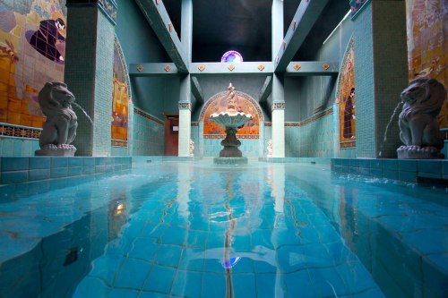 A serene indoor pool with turquoise tiles, a central fountain, and decorative arches, illuminated by soft light.