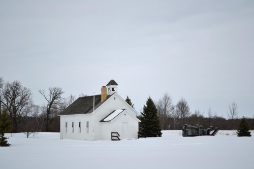 A small white schoolhouse surrounded by snow, with trees and a plow in the background under a cloudy sky.