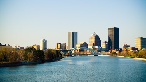 A city skyline with modern buildings along a river, featuring a bridge and clear blue sky.
