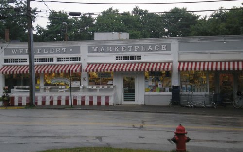 A storefront of Wellfleet Marketplace with striped awnings, surrounded by trees and a wet street.