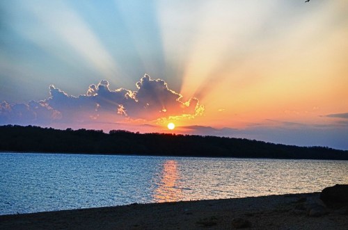 A serene sunset over a calm lake, with vibrant rays of light breaking through clouds and reflecting on the water.