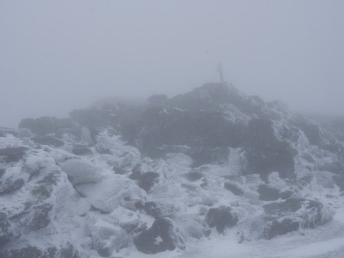 A rocky landscape shrouded in fog and snow, with a faint outline of a cross in the distance.