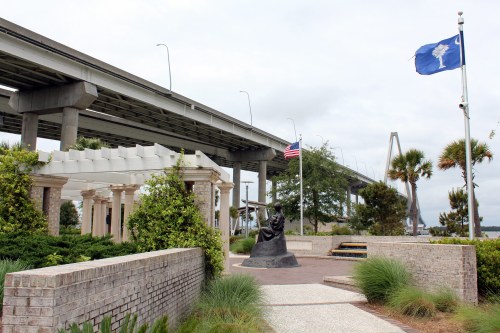 A park scene featuring a statue, flags, and a bridge overhead, surrounded by greenery and brick pathways.