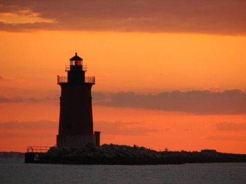 Silhouette of a lighthouse against a vibrant orange sunset over the water.