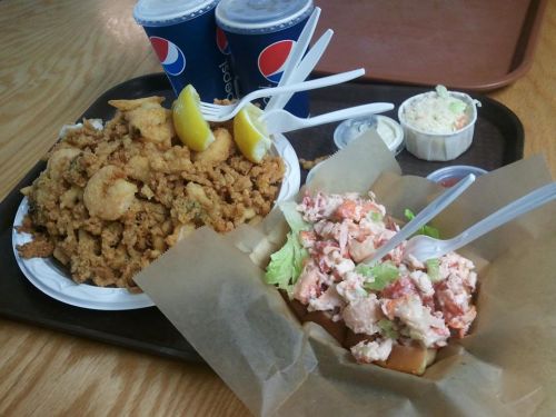 A tray with fried seafood, lemon wedges, a lobster roll, coleslaw, and two cups of soda.