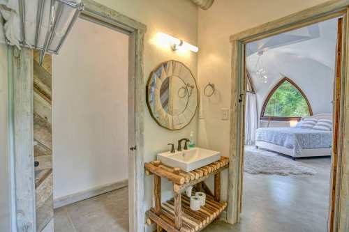 A rustic bathroom with a wooden sink stand, mirror, and an arched doorway leading to a cozy bedroom.