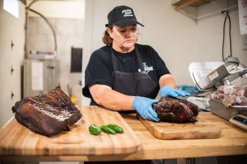 A woman in a black apron prepares smoked meat on a wooden cutting board, with jalapeños nearby.