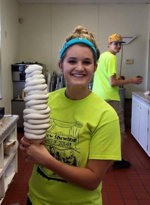 A smiling young woman in a bright green shirt holds a large swirl ice cream cone, with a person in the background.