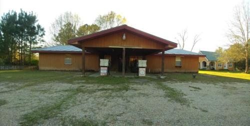 A wooden building with a sloped roof, surrounded by trees and a gravel area, under a clear sky.