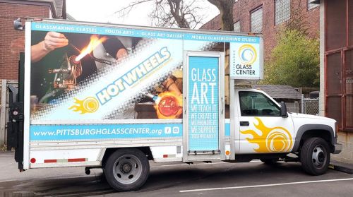 A mobile glass art studio truck featuring "Hot Wheels" and promoting glassmaking classes and youth arts.