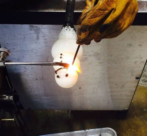 A gloved hand shapes molten glass with a tool, creating a glowing glass object in a workshop setting.