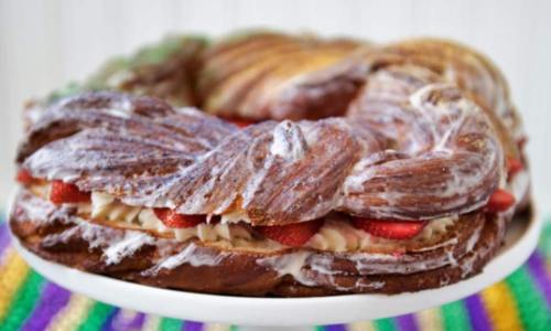 A colorful braided cake topped with icing and filled with strawberries, displayed on a white cake stand.