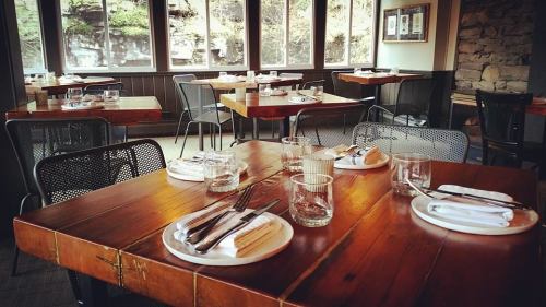 Cozy restaurant interior with wooden tables, glassware, and silverware, surrounded by large windows.