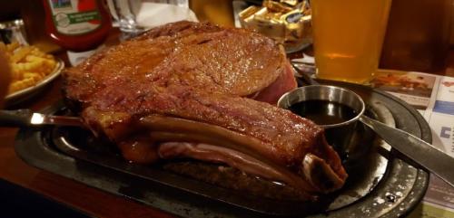 A large, juicy prime rib served on a sizzling platter with a side of sauce, accompanied by fries and a pint of beer.