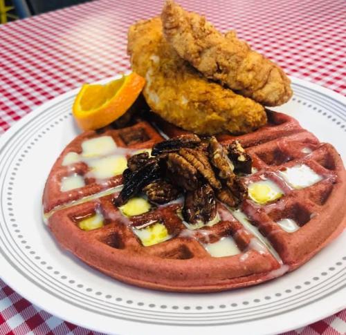 A plate featuring red velvet waffles topped with butter, pecans, and two pieces of fried chicken, garnished with an orange slice.