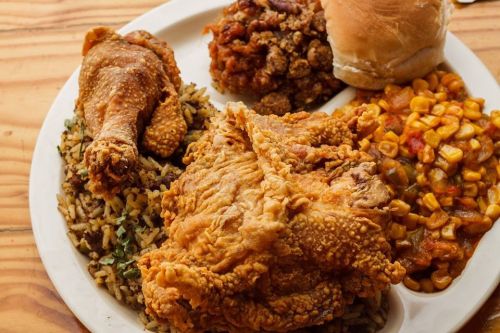 A plate of fried chicken, rice, corn, baked beans, and a bun on a wooden table.