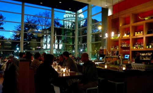 A cozy restaurant interior with patrons, large windows showcasing a cityscape at dusk. Warm lighting creates an inviting atmosphere.