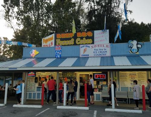 A colorful roadside eatery with a line of customers outside, featuring signs for BBQ, ice cream, and funnel cakes.