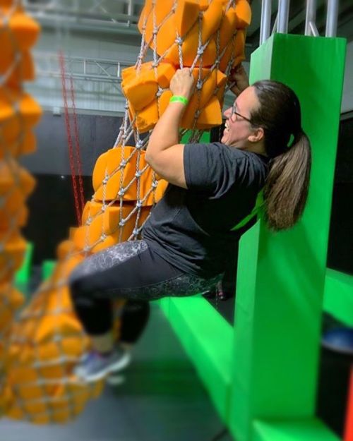 A person climbs a rope structure surrounded by orange foam blocks in a vibrant indoor activity space.