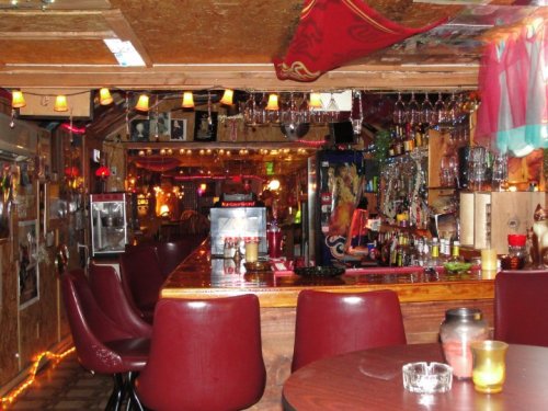 Cozy bar interior with wooden counter, red chairs, colorful lights, and various decorations on the walls.