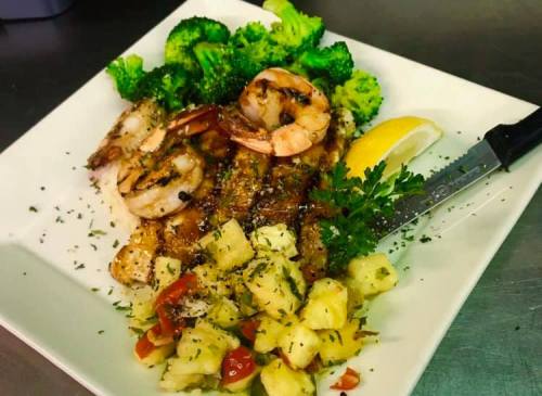 A plate of grilled shrimp and fish with broccoli, lemon, and a side of diced pineapple and tomato salad.