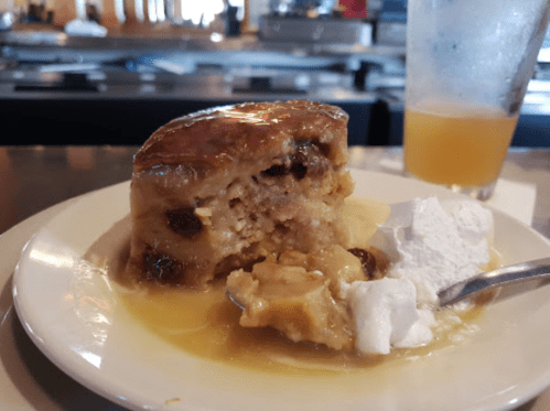 A slice of bread pudding drizzled with sauce, served with whipped cream on a plate, with a drink in the background.