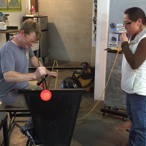 A glassblower shapes molten glass while a woman observes, both engaged in the art of glassblowing in a studio.