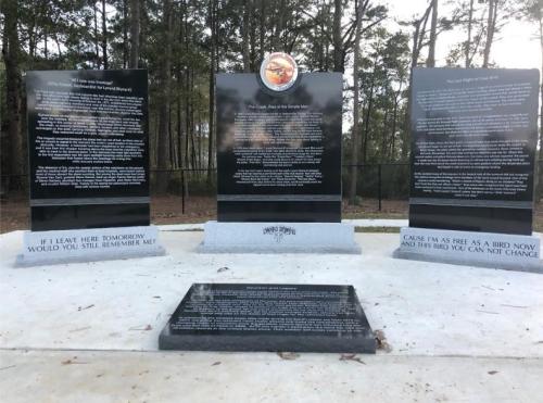 Three black memorial plaques with inscriptions, surrounded by trees, honoring service and remembrance.