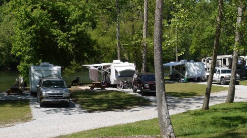 A scenic campsite with RVs and trucks parked near a river, surrounded by trees and green grass.