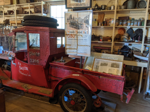A vintage red truck with a sign for Woods Model No. 5, surrounded by various antique items in a museum display.