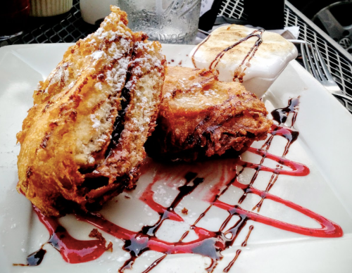 Fried dessert with powdered sugar, chocolate drizzle, and a side of dipping sauce on a white plate.