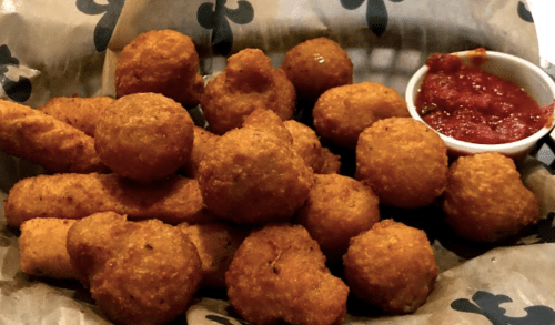 A basket of golden-brown fried snacks, including round and stick shapes, served with a small cup of marinara sauce.