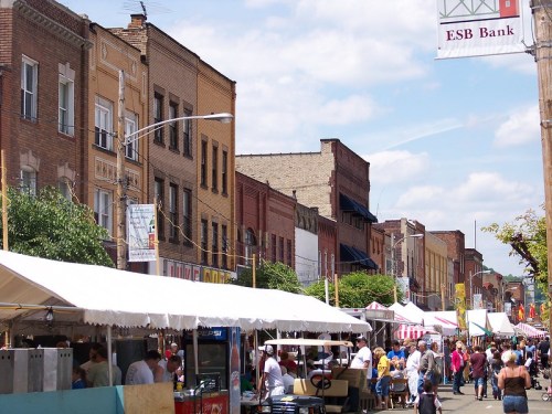 A bustling street fair with vendors and colorful tents, surrounded by historic brick buildings under a clear blue sky.