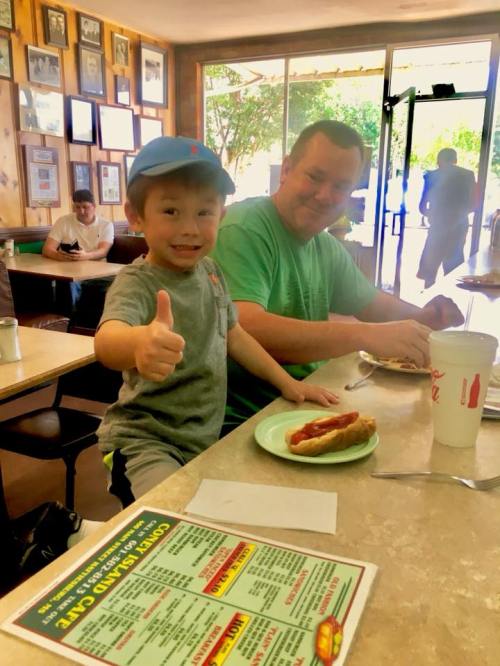 A young boy gives a thumbs up while sitting at a table with a man, both enjoying hot dogs in a casual diner setting.
