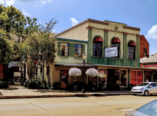 Historic building with green and yellow facade, featuring large windows and hanging flower baskets, on a sunny street.