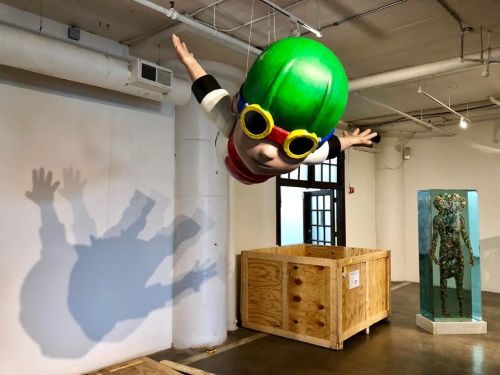 A large, colorful sculpture of a child with green hair and sunglasses appears to fly above a wooden crate in an art gallery.