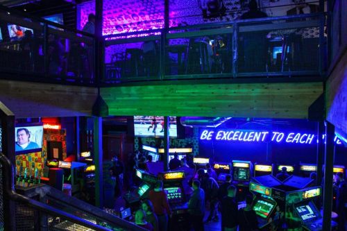 A vibrant arcade bar with neon lights, vintage arcade games, and people socializing on multiple levels.