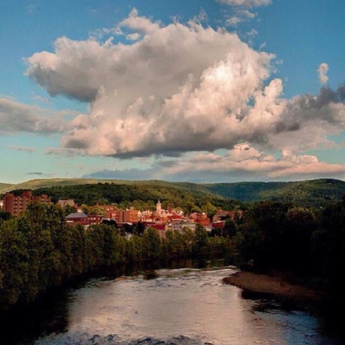 A scenic view of a river flowing through a town, surrounded by green hills and a blue sky with fluffy clouds.
