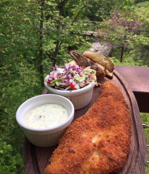 A wooden platter with crispy fish, coleslaw, and potato wedges, set against a lush green background.