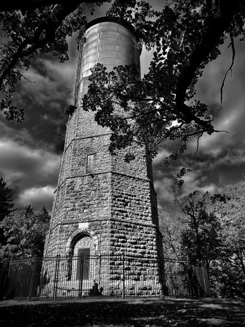 A tall stone water tower surrounded by trees, set against a dramatic cloudy sky in black and white.