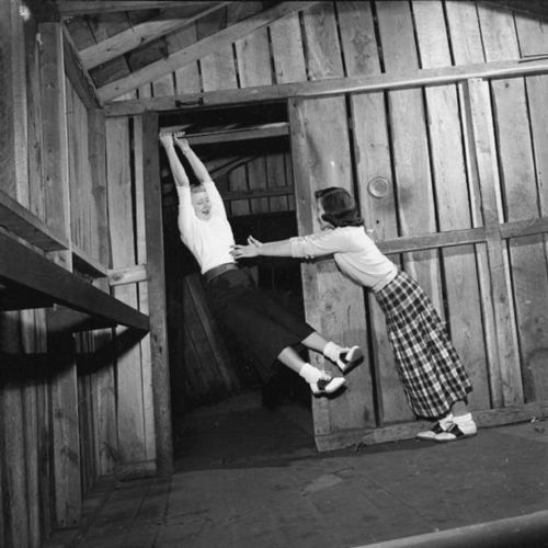 Two women playfully interacting in a rustic wooden room, one hanging from a doorframe while the other supports her.