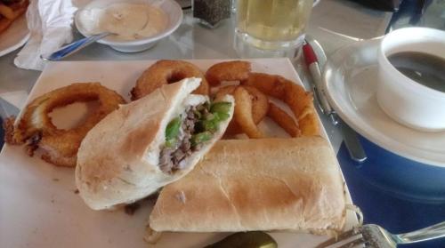 A plate with a sandwich filled with beef and peppers, onion rings, a small bowl of dip, and a cup of coffee.