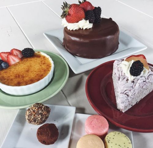 A variety of desserts including a chocolate cake, berry cheesecake, crème brûlée, and colorful macarons on plates.