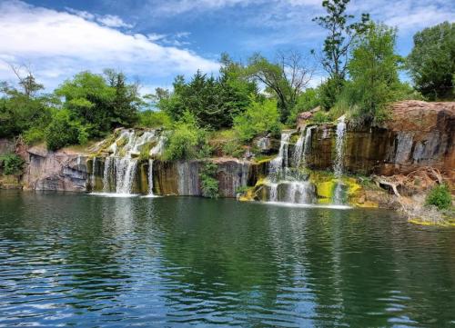 A serene waterfall cascading over rocky cliffs into a calm, greenish-blue lake surrounded by lush trees and blue skies.