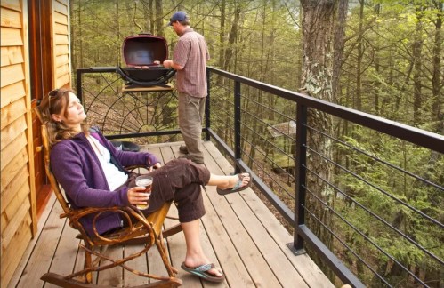 A woman relaxes on a deck with a drink while a man grills nearby in a forested setting.