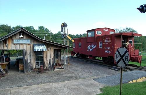 A vintage red caboose next to a rustic train station, with a railroad crossing sign and green trees in the background.