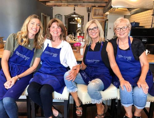 Four women in blue aprons smile while sitting together in a kitchen setting, showcasing a warm and friendly atmosphere.