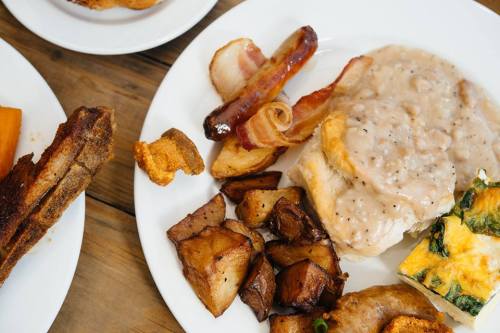 A plate of hearty breakfast featuring biscuits with gravy, bacon, sausage, roasted potatoes, and a vegetable frittata.